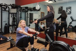 A fitness trainer guides a woman on a rowing machine, while others follow a workout in the background of a gym.
