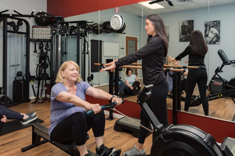 A fitness trainer guides a woman on a rowing machine, while others follow a workout in the background of a gym.