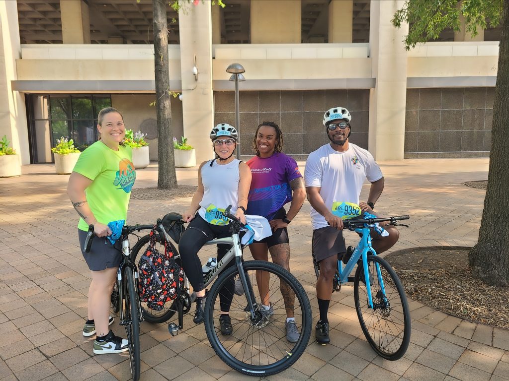 Four cyclists in athletic gear pose with their bikes, smiling outdoors near a building, promoting heart health through movement.