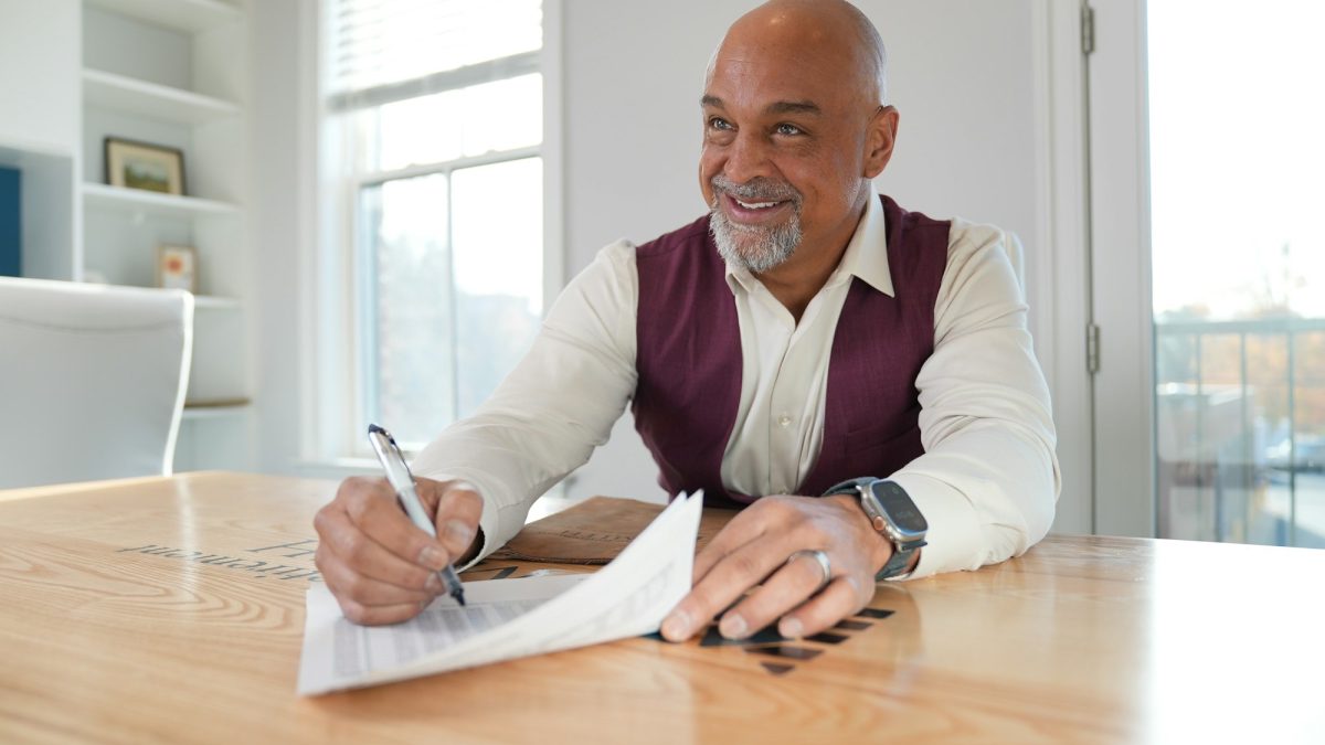 Man in a burgundy vest smiles while writing on a document at a wooden table in a bright room.