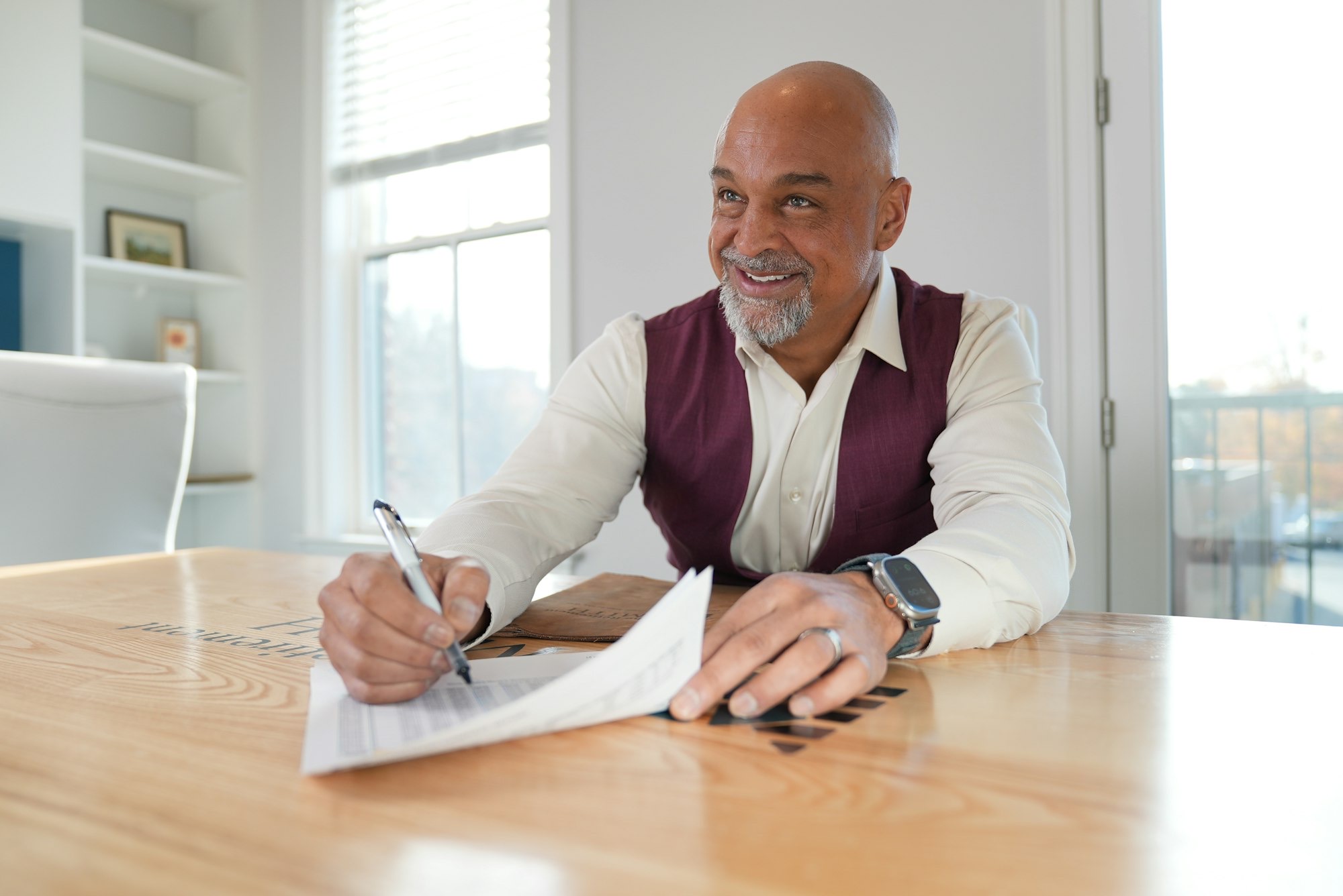 Man in a burgundy vest smiles while writing on a document at a wooden table in a bright room.