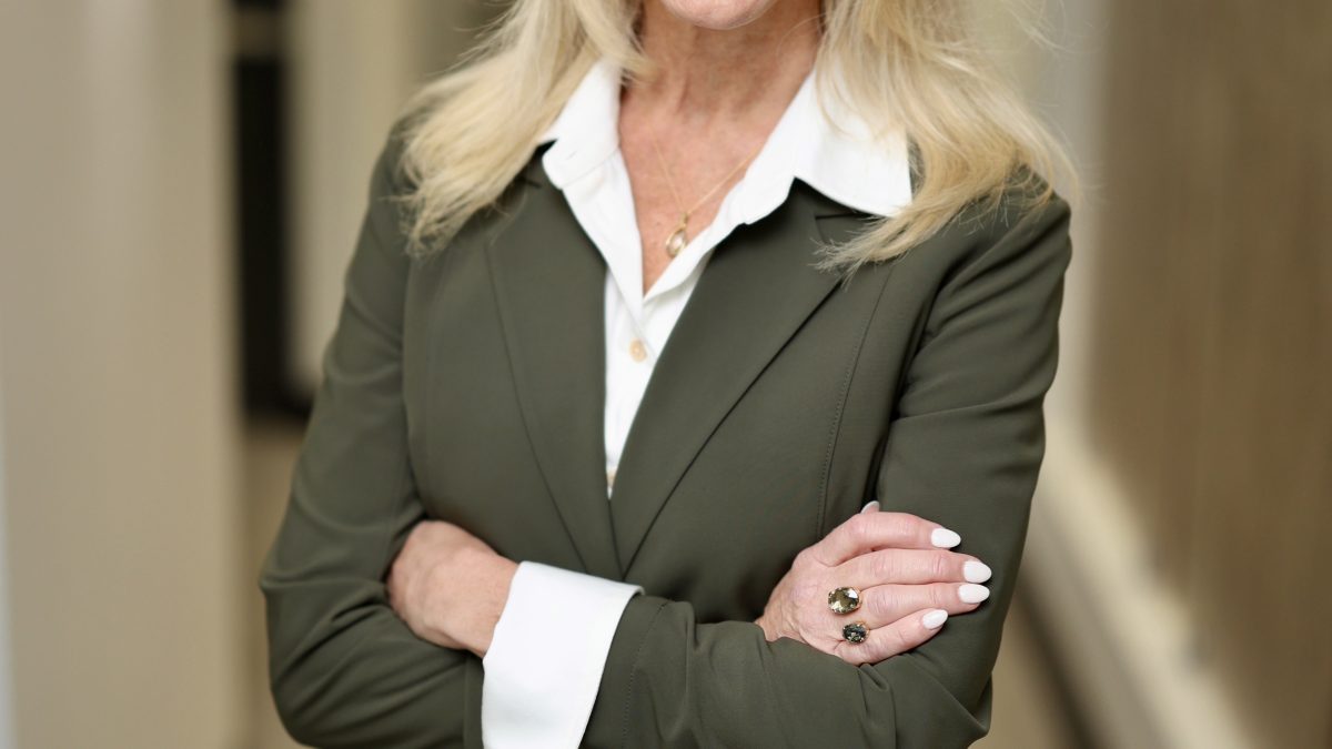 Smiling woman with long blonde hair, wearing a green blazer and white blouse, stands confidently with arms crossed in a hallway.