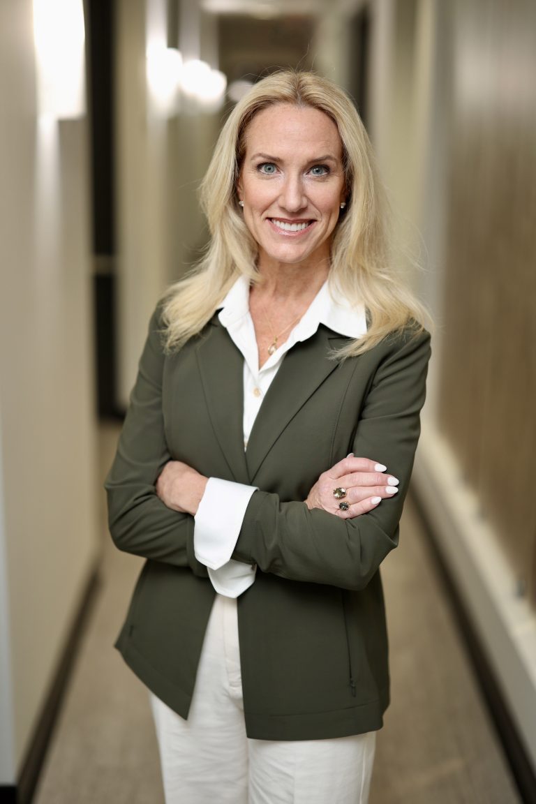 Smiling woman with long blonde hair, wearing a green blazer and white blouse, stands confidently with arms crossed in a hallway.