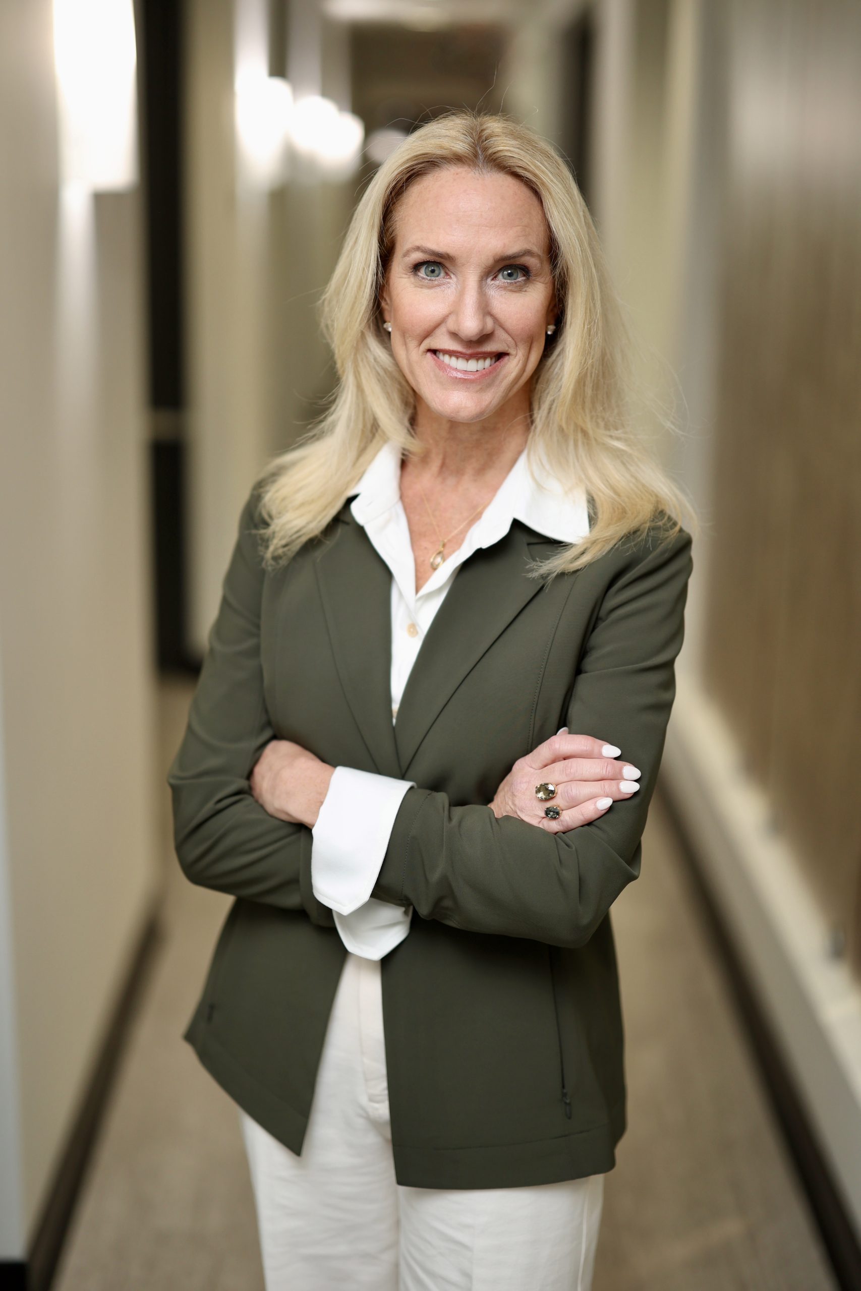 Smiling woman with long blonde hair, wearing a green blazer and white blouse, stands confidently with arms crossed in a hallway.