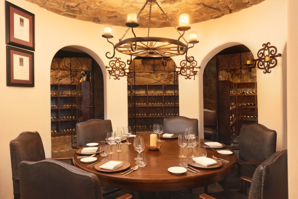 Elegant dining room featuring a round wooden table, surrounded by plush chairs and adorned with wine racks and decorative lighting.