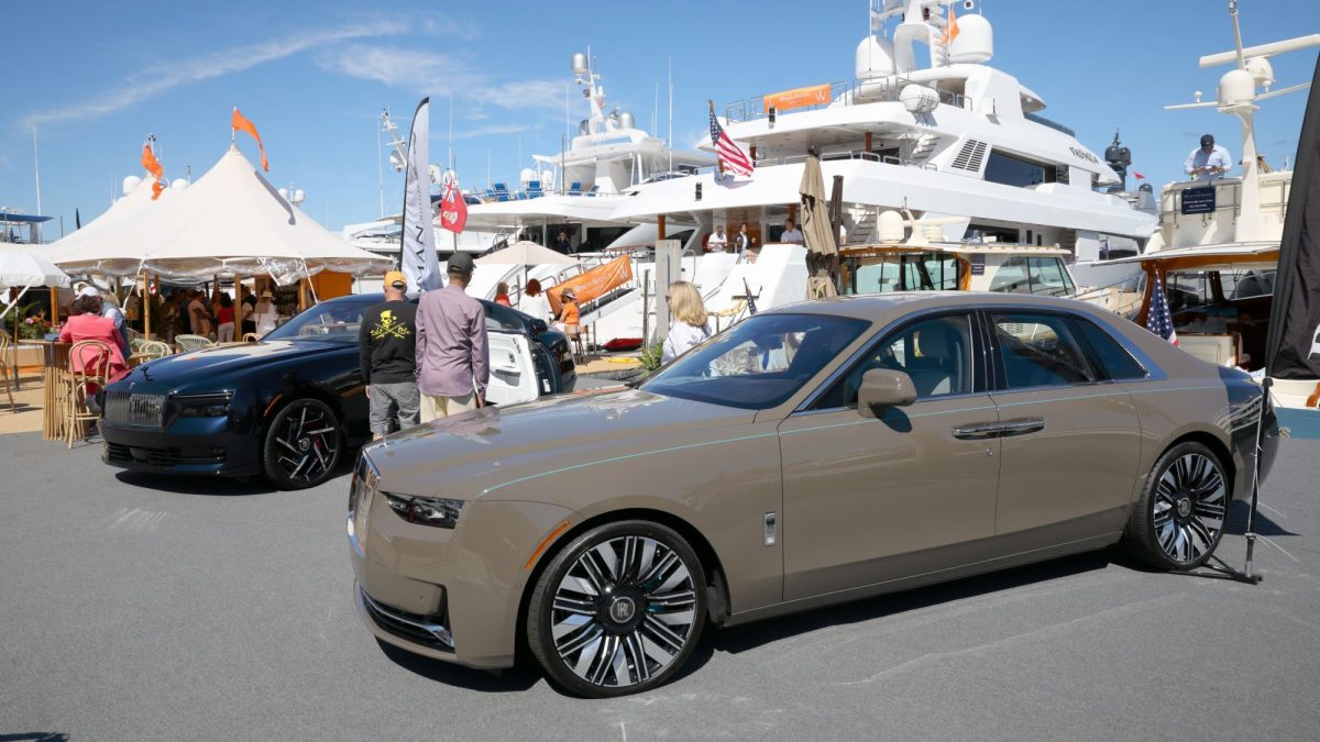 Luxury cars on display at the Palm Beach International Boat Show with yachts in the background and attendees exploring the event.