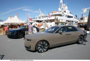 Luxury cars on display at the Palm Beach International Boat Show with yachts in the background and attendees exploring the event.