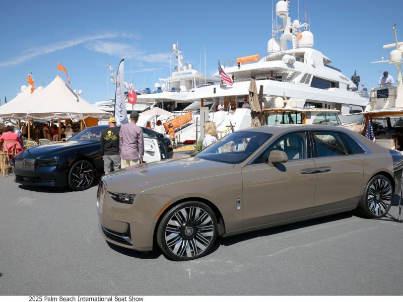 Luxury cars on display at the Palm Beach International Boat Show with yachts in the background and attendees exploring the event.