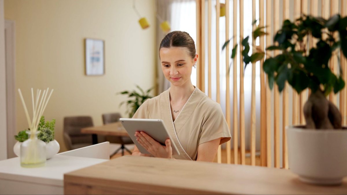 A spa professional uses a tablet at a reception desk, surrounded by greenery and modern decor.