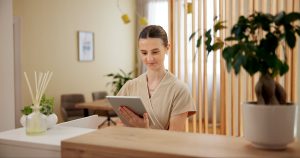 A spa professional uses a tablet at a reception desk, surrounded by greenery and modern decor.