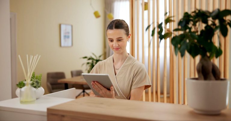 A spa professional uses a tablet at a reception desk, surrounded by greenery and modern decor.