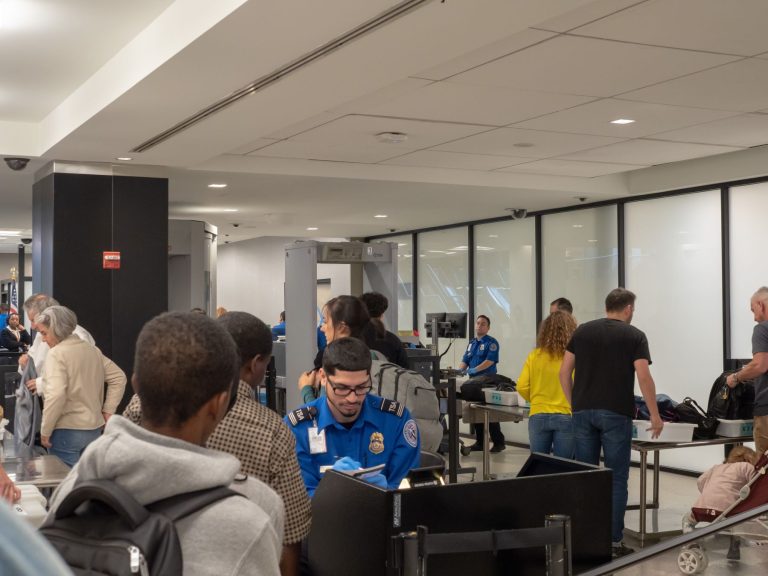 Travelers navigate a busy airport security checkpoint with TSA agents and luggage screening in view.