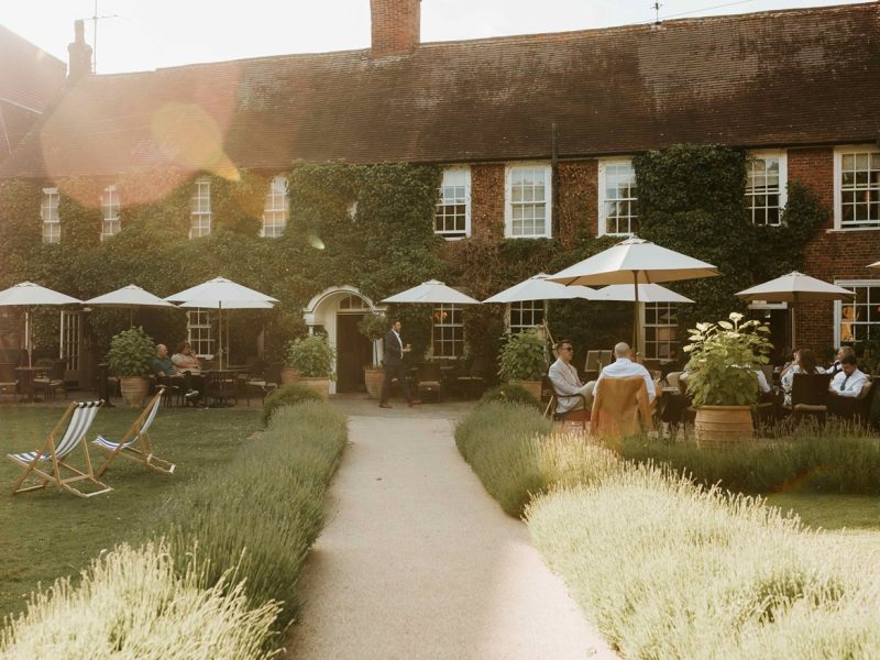 Guests enjoy outdoor seating at The Bush Farnham, surrounded by greenery and patio umbrellas on a sunny day.