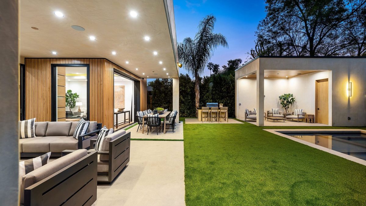 Modern outdoor living space with seating, dining area, and ambient lighting, surrounded by greenery at dusk.