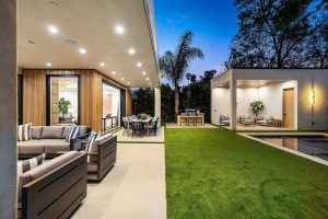 Modern outdoor living space with seating, dining area, and ambient lighting, surrounded by greenery at dusk.