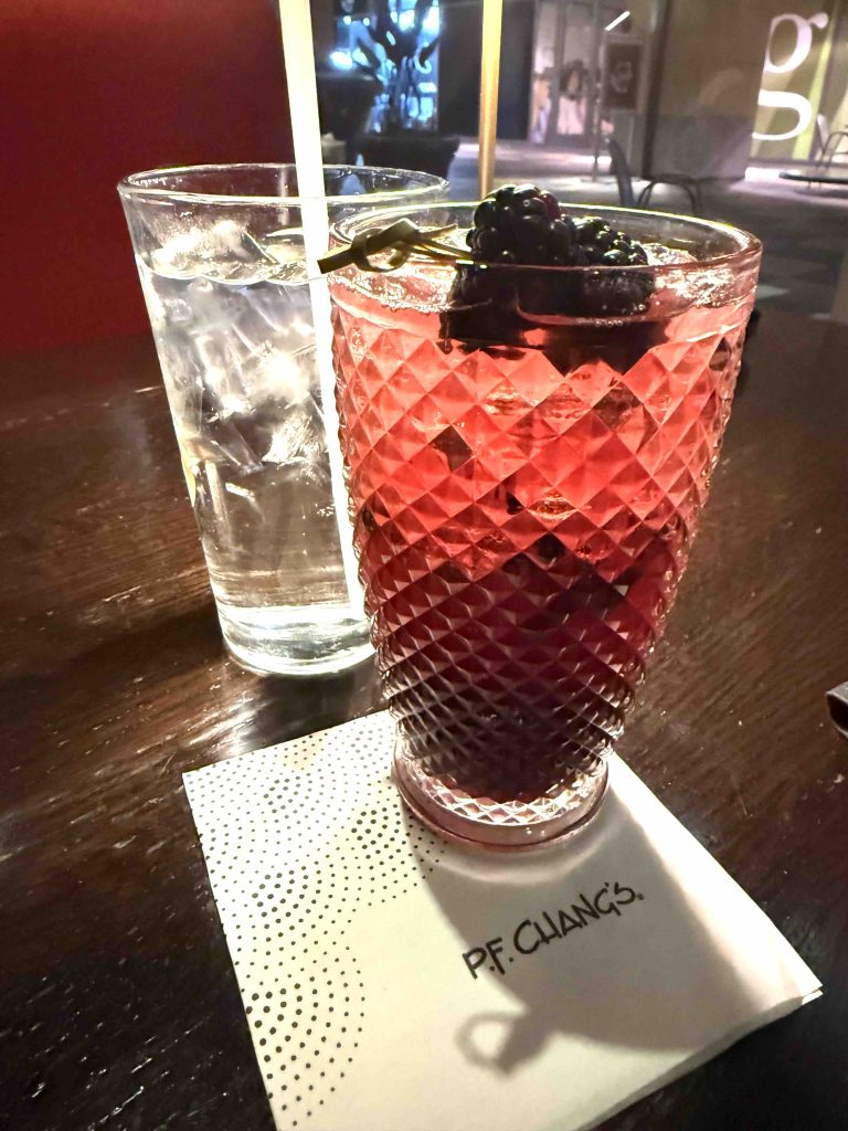 A colorful cocktail garnished with blackberries next to a glass of water on a P.F. Chang's branded napkin.