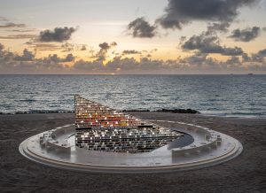 A large, triangular sculpture resembling a book display is set on the beach at sunset, reflecting the ocean and sky.