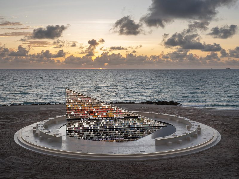 A large, triangular sculpture resembling a book display is set on the beach at sunset, reflecting the ocean and sky.
