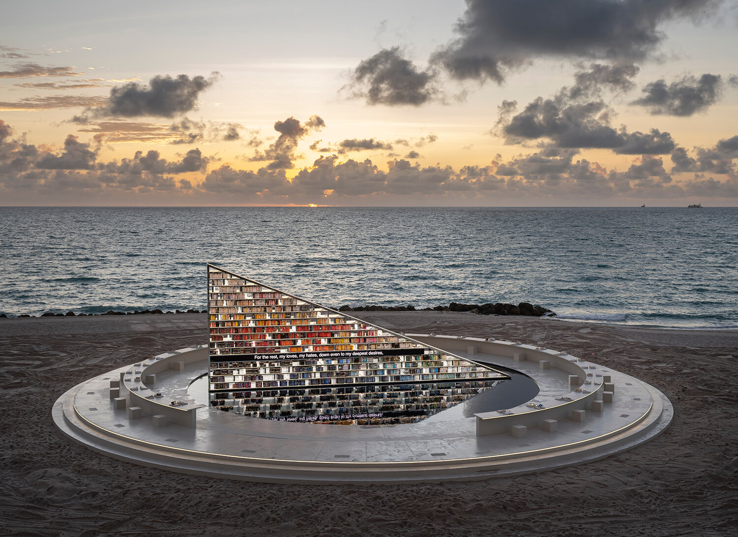 A large, triangular sculpture resembling a book display is set on the beach at sunset, reflecting the ocean and sky.