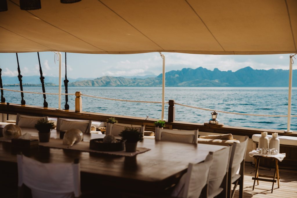 Luxury yacht deck with a wooden dining table overlooking calm waters and distant hills in Indonesia.