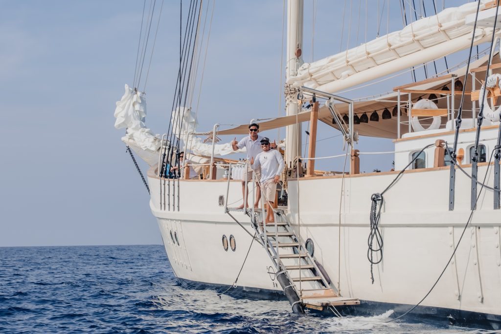 Two crew members smiling and waving from the deck of a luxury sailing yacht in the ocean, showcasing Indonesian beauty.
