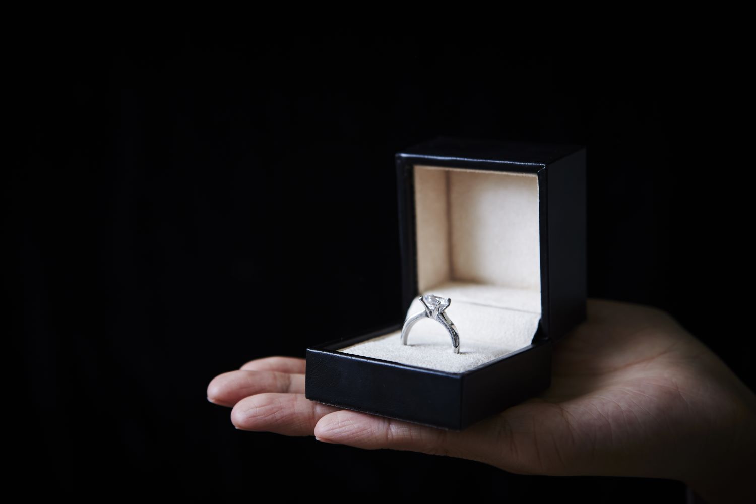A silver engagement ring displayed in an open black box, held in a hand against a dark background.