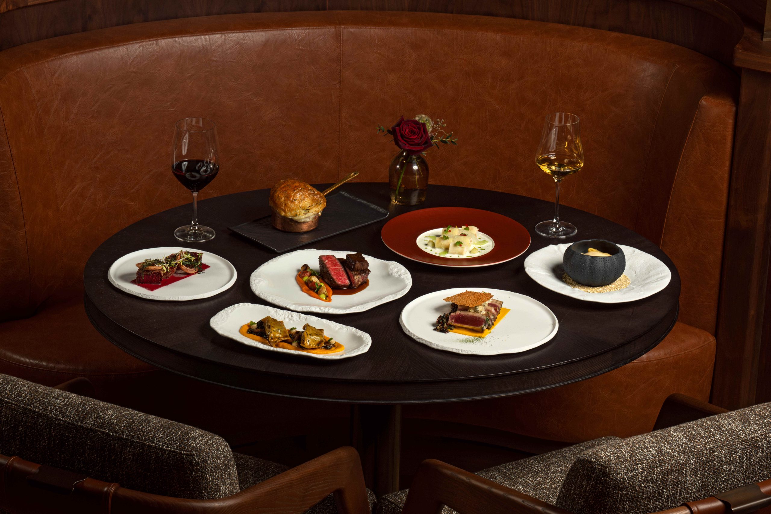 A round table set with various gourmet dishes, wine glasses, and a rose in a vase, highlighting a luxurious dining experience.