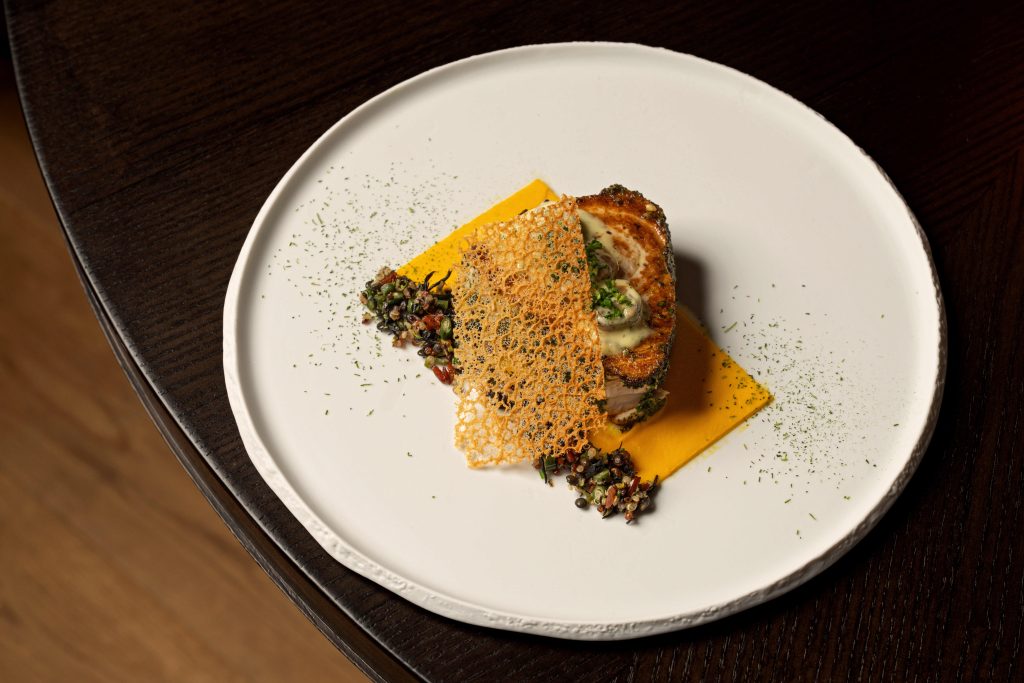 Sautéed sustainable fish served with a vibrant yellow puree and colorful grain medley on a white plate.