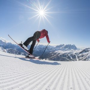 Skier in a red jacket gliding down a snow-covered slope with a panoramic view of the Dolomites under a bright sun.