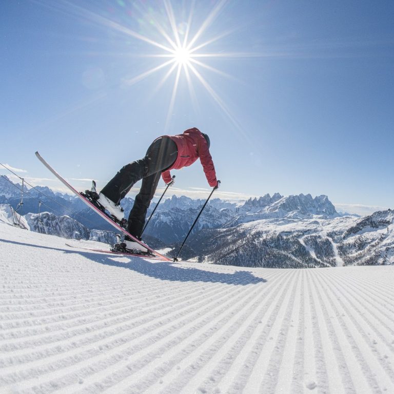Skier in a red jacket gliding down a snow-covered slope with a panoramic view of the Dolomites under a bright sun.