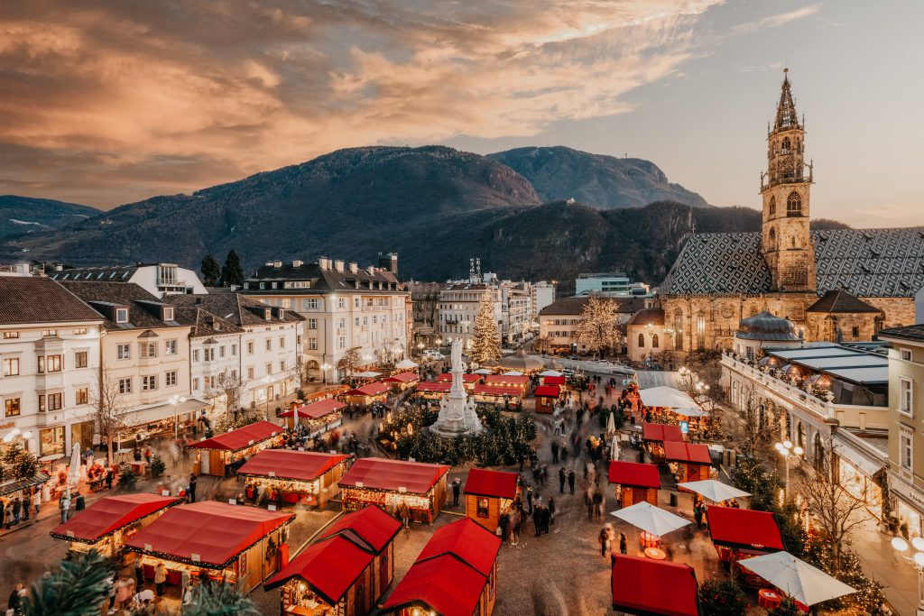 Christmas market with decorated stalls and crowds, set against a mountainous backdrop at sunset.