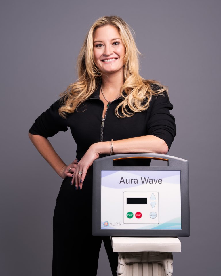 Woman with long blonde hair smiles while standing next to an Aura Wave device, promoting accessible pediatric care services.