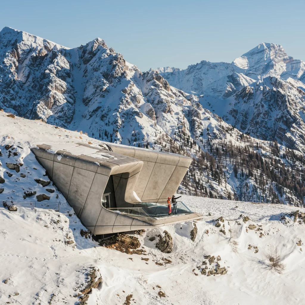 Modern architectural viewpoint in the snowy Dolomites with skiers enjoying the panoramic mountain scenery.
