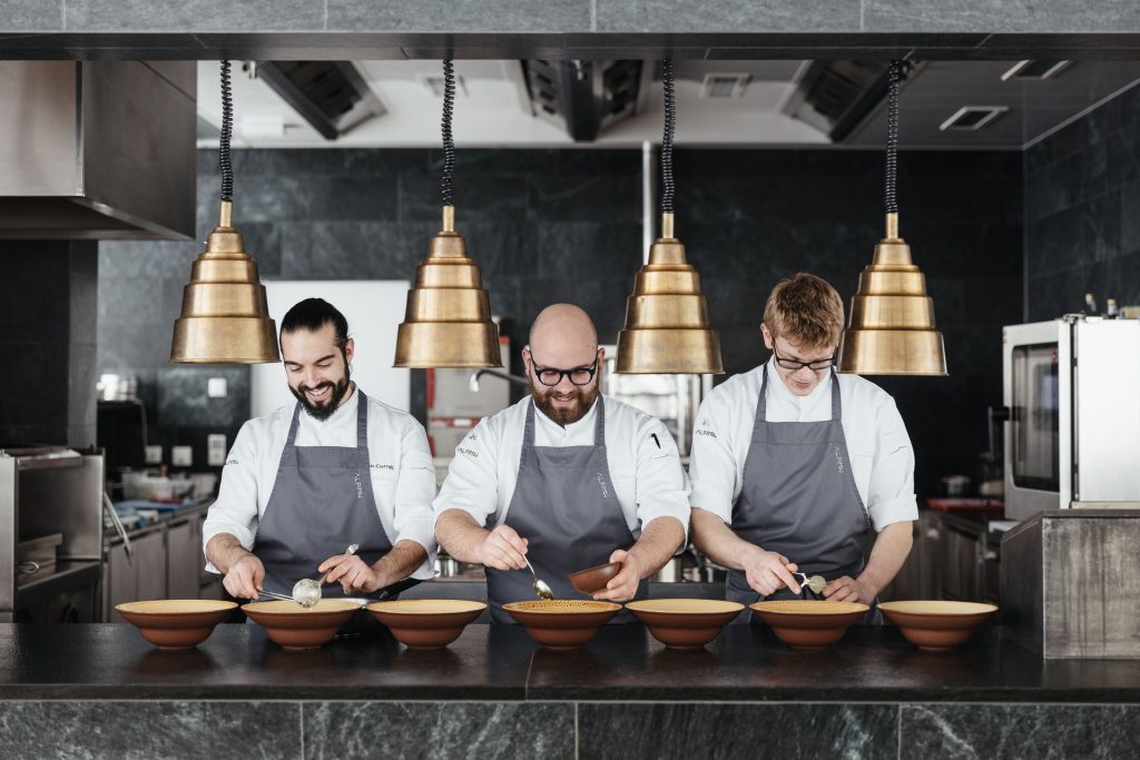 Three chefs in a modern kitchen prepare gourmet dishes, focusing on plating in front of elegant bowls.