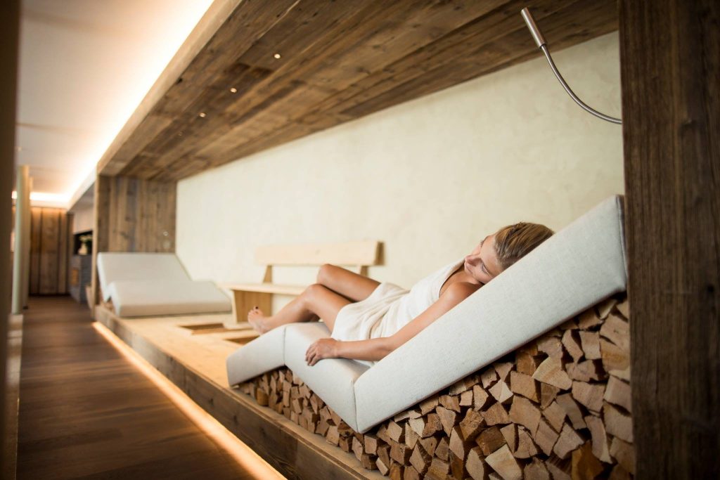 A woman relaxes on a contemporary lounge chair made of wood and fabric in a spa-like room with soft lighting.