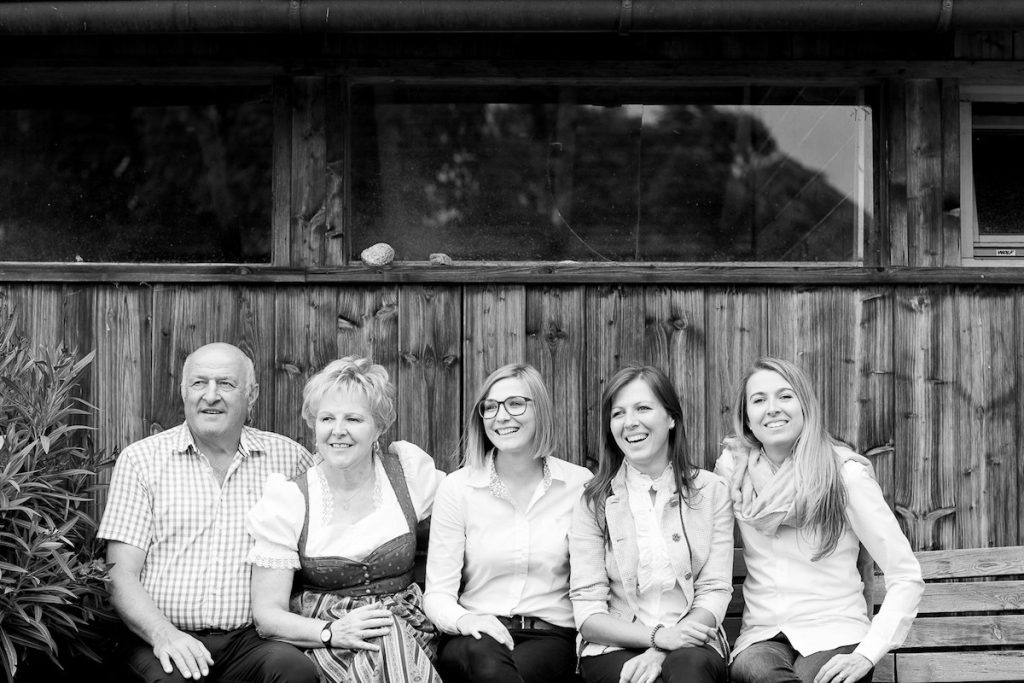 Group of five people smiling and sitting on a bench in front of a wooden wall, dressed in casual and traditional attire.