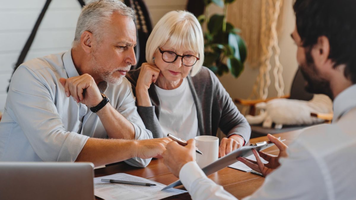Couple discusses estate planning with a financial advisor, reviewing documents at a wooden table.