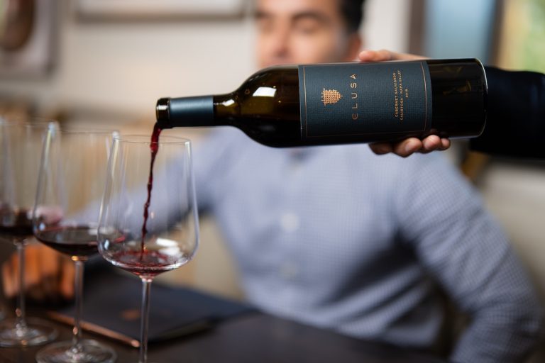 Red wine being poured from an Elusa bottle into glasses during a tasting session at Elusa Winery.