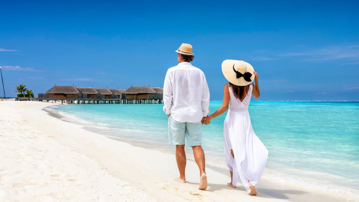 A couple strolls hand in hand along a serene beach, with clear turquoise water and overwater bungalows in the background.