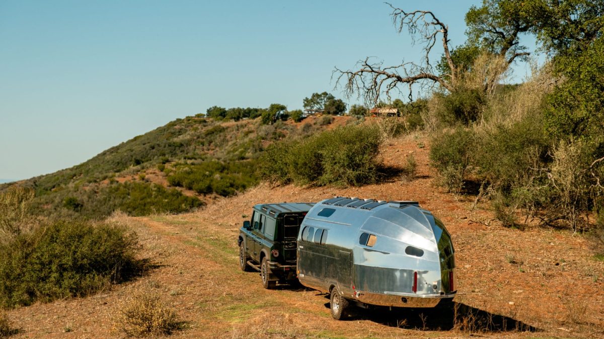 A shiny silver RV trailer is parked beside a dark SUV on a dirt path surrounded by hills and sparse vegetation.