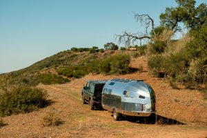 A shiny silver RV trailer is parked beside a dark SUV on a dirt path surrounded by hills and sparse vegetation.