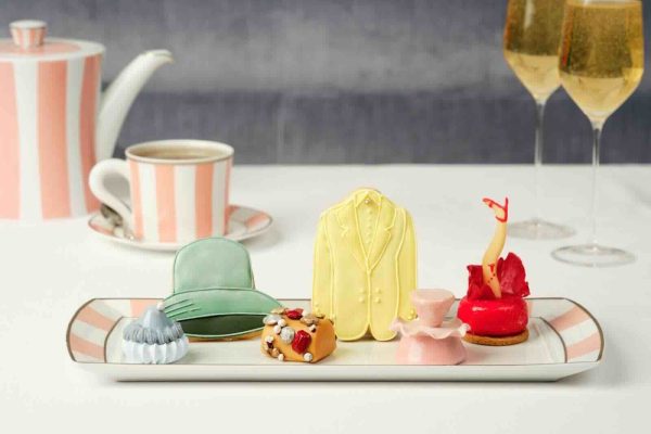 Colorful pastries resembling fashion items are displayed on a tray, alongside tea and sparkling drinks in an elegant setting.