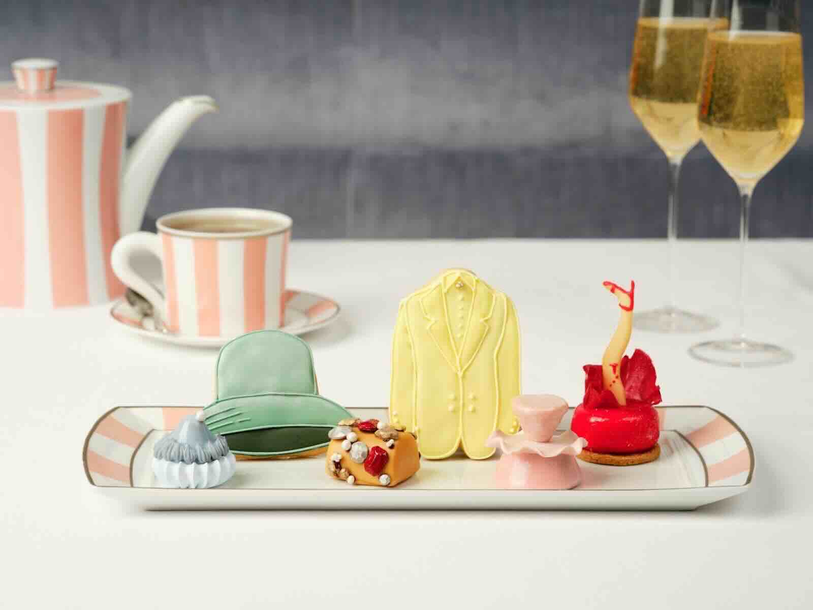 Colorful pastries resembling fashion items are displayed on a tray, alongside tea and sparkling drinks in an elegant setting.