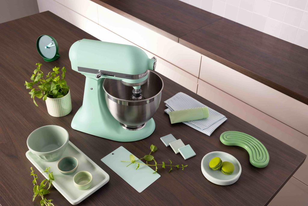 Spearmint KitchenAid mixer on a wooden countertop with green accessories, bowls, and a plant.