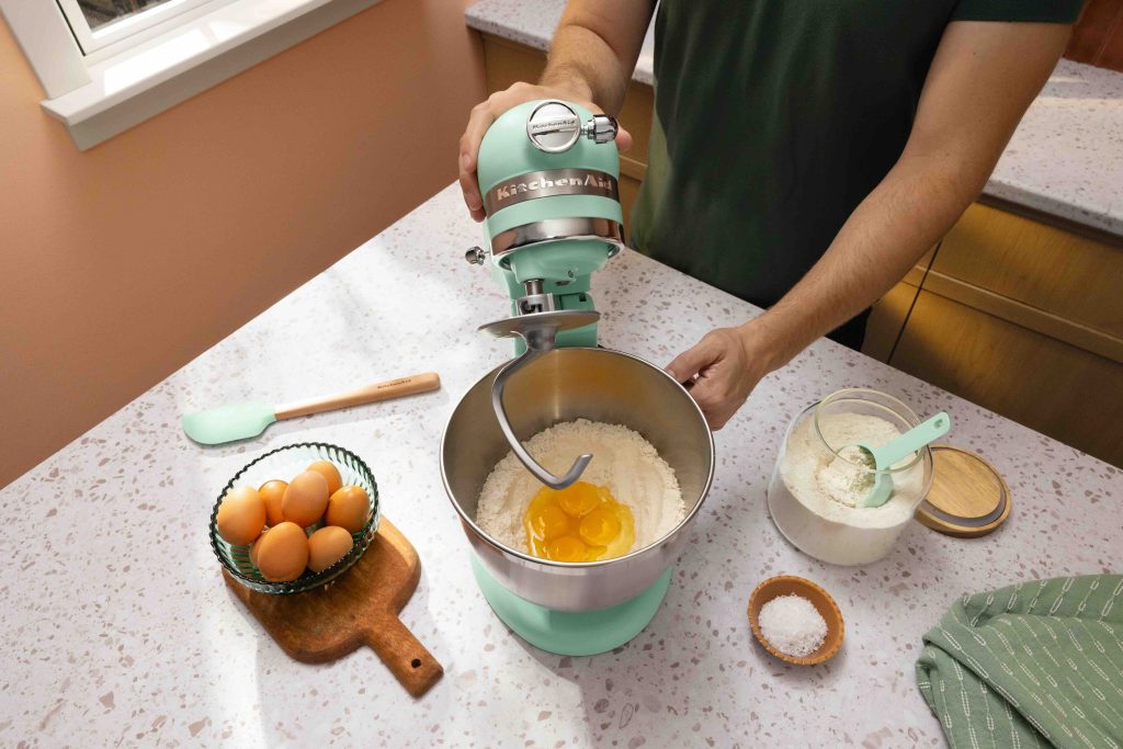 A KitchenAid spearmint mixer is shown in use, mixing flour and eggs, with eggs and salt nearby on a countertop.