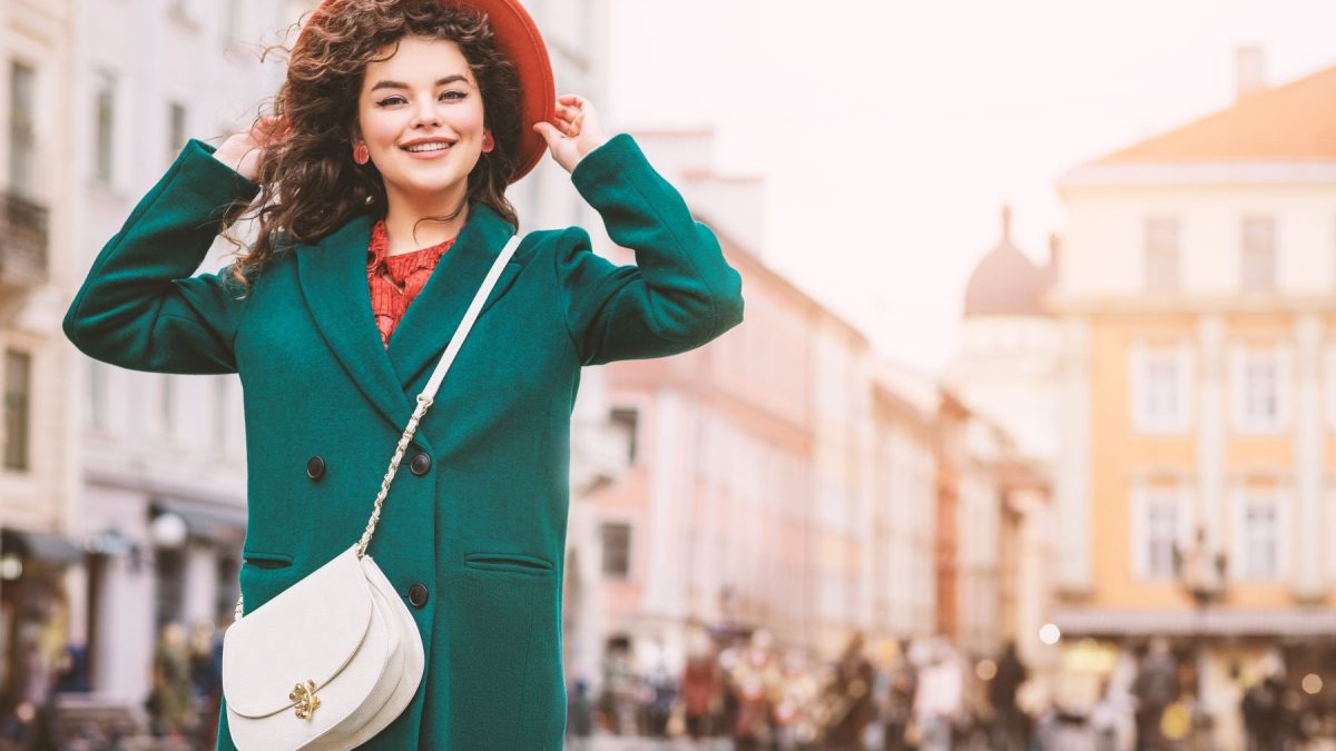 A woman wearing a green coat and red hat smiles while holding her hat, showcasing a white crossbody-to-clutch hybrid bag.