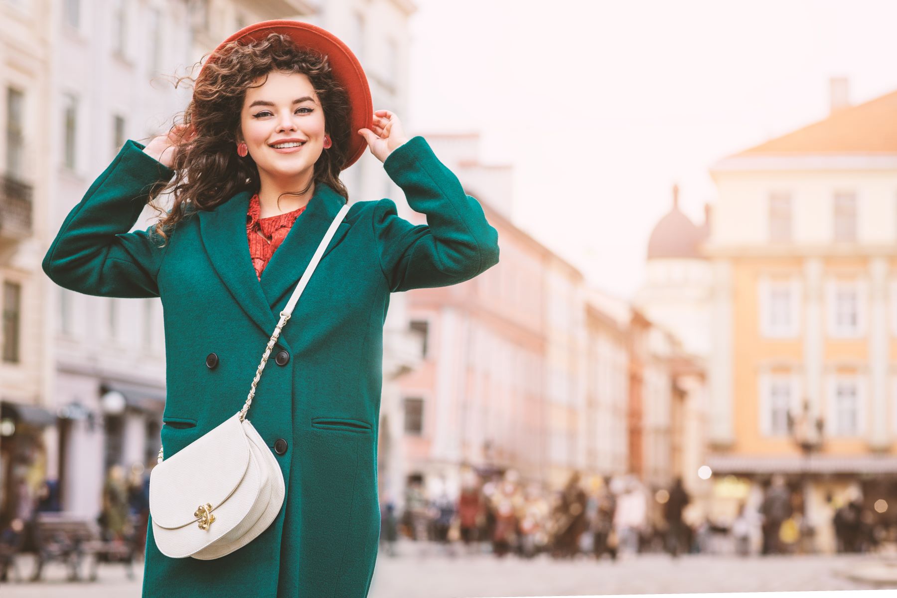 A woman wearing a green coat and red hat smiles while holding her hat, showcasing a white crossbody-to-clutch hybrid bag.