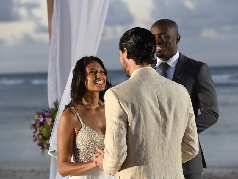 A couple exchanges vows on a beach during their wedding ceremony, with a celebrant and ocean backdrop.