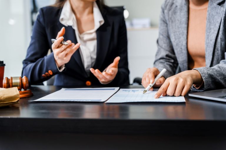Two women discussing documents at a table, with a gavel and coffee cup nearby, focused on legal matters.