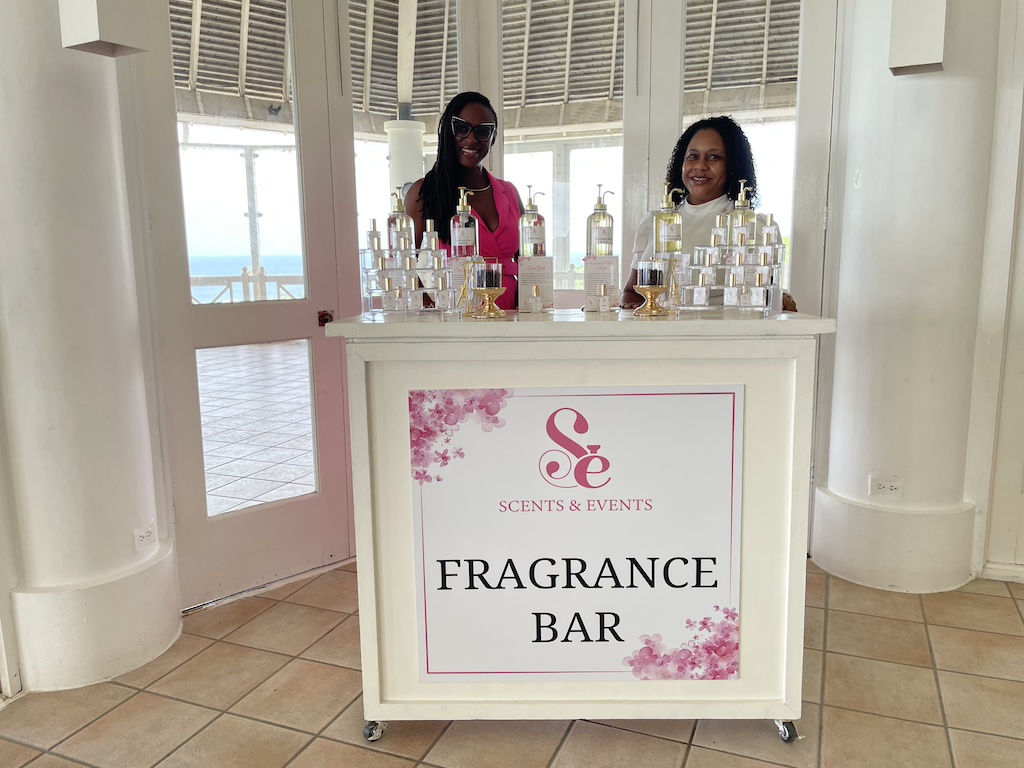 Two women stand behind a fragrance bar with various bottles displayed, promoting scents and events in a bright setting.